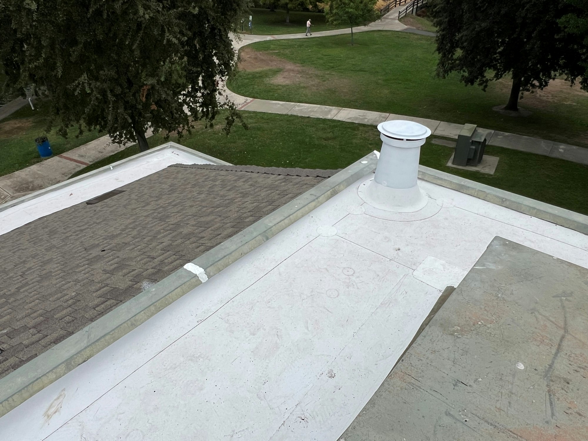 A rooftop with a white vent, overlooking a park with trees, paths, and a person walking.