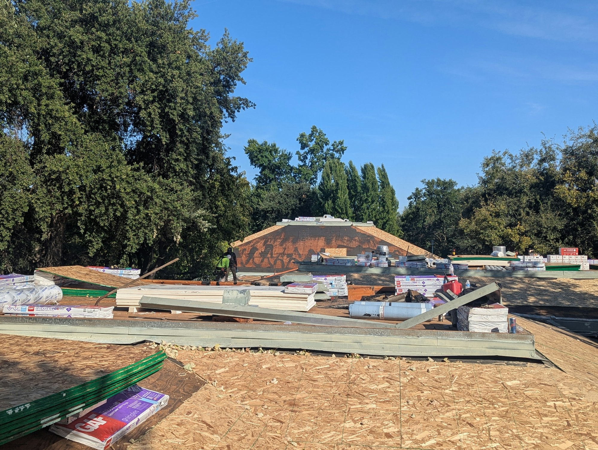 Roof under construction with materials scattered, trees in the background, and a clear blue sky overhead.