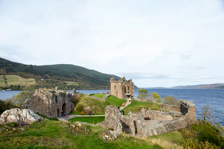 A scenic view of a castle ruin beside a lake, surrounded by green hills, with visitors exploring the site.
