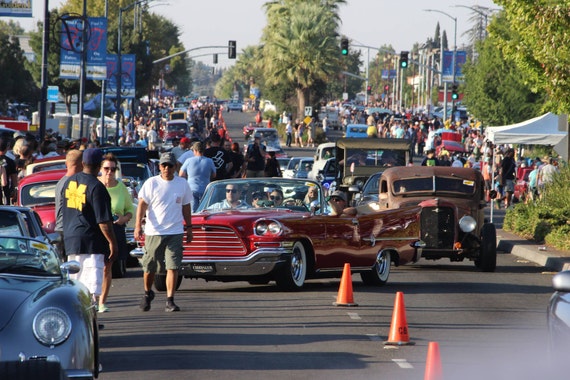 A street parade with classic cars, people walking, and orange cones marking the route.