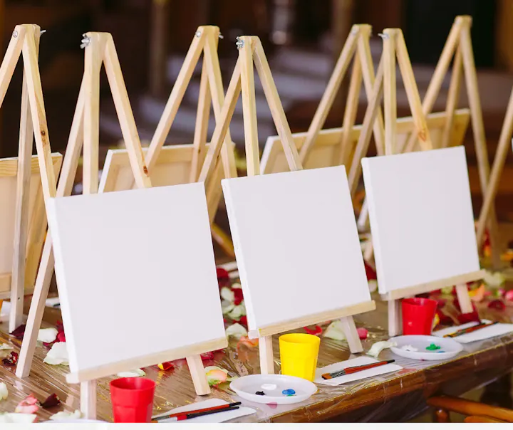 Blank canvases on wooden easels with paint palettes, brushes, and colorful cups are arranged on a table.