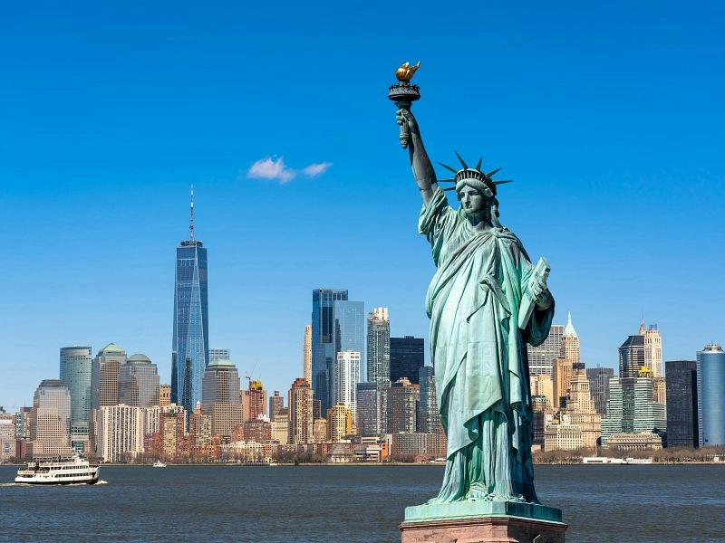 The image shows the Statue of Liberty standing tall with the New York City skyline in the background on a clear day.