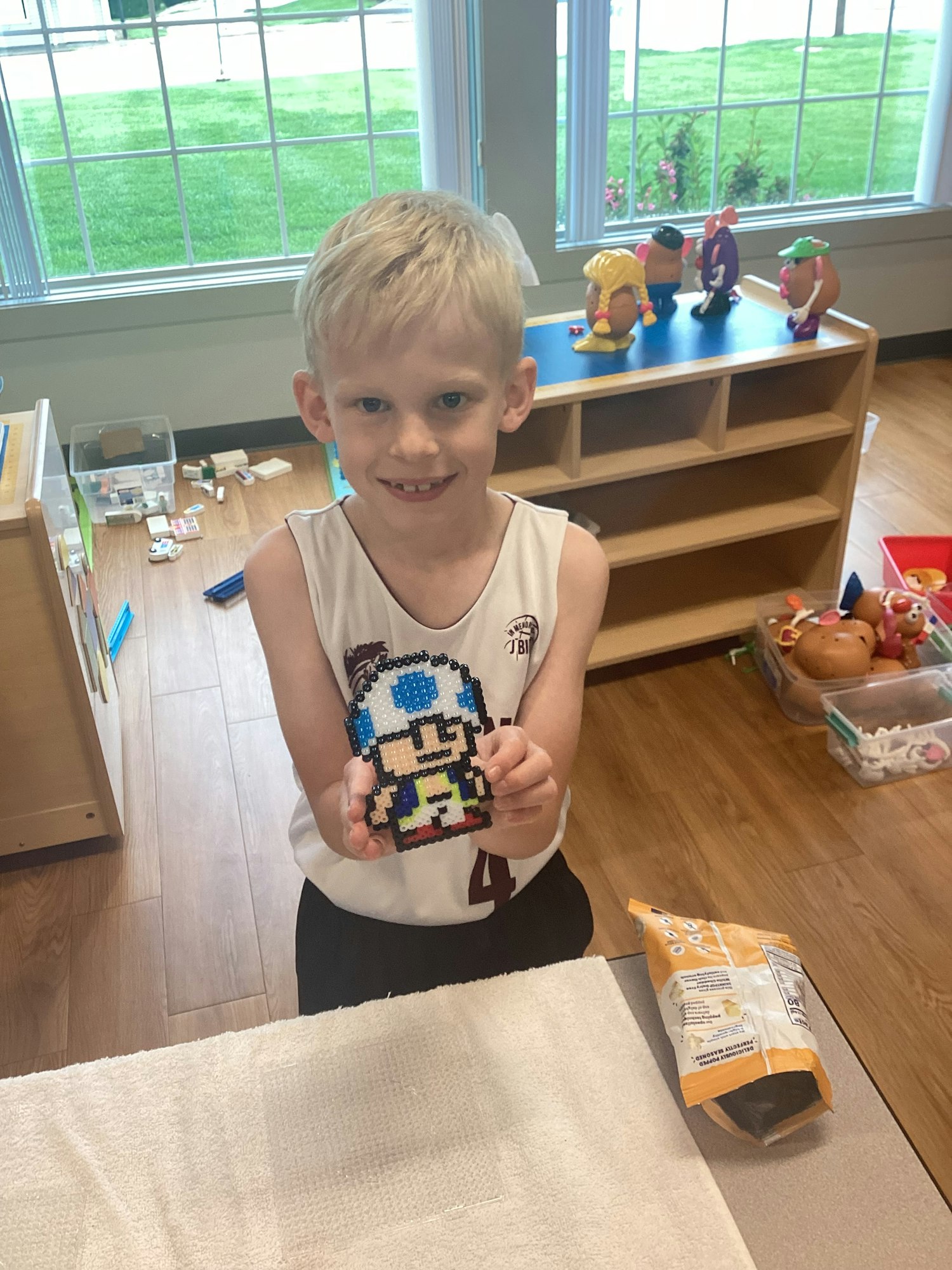 Child holding a pixel art creation, with toys and an open snack bag on a table in a playroom with wooden floors.