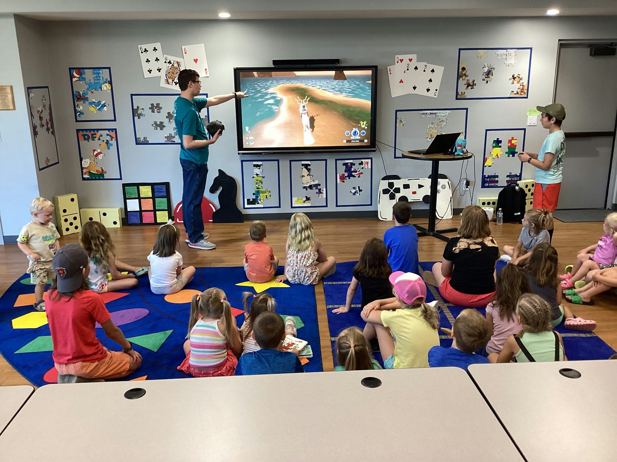 Children watching a presentation in a classroom with colorful decor and a screen showing a game.