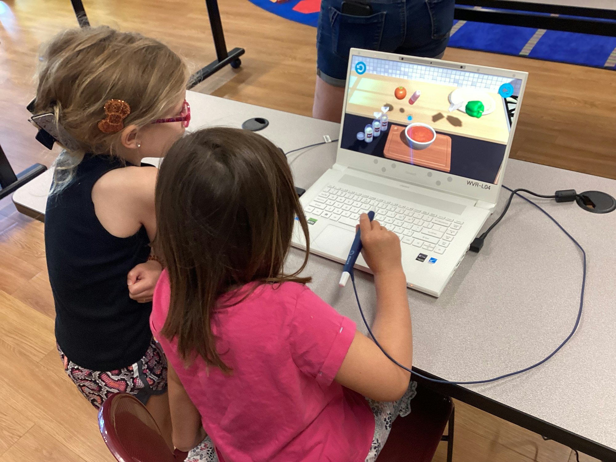 Two children using a laptop with a stylus, interacting with a cooking simulation game on the screen.