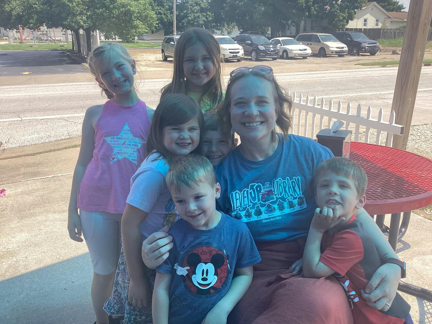 A group of smiling children and a woman sitting outdoors near a red table and white fence.