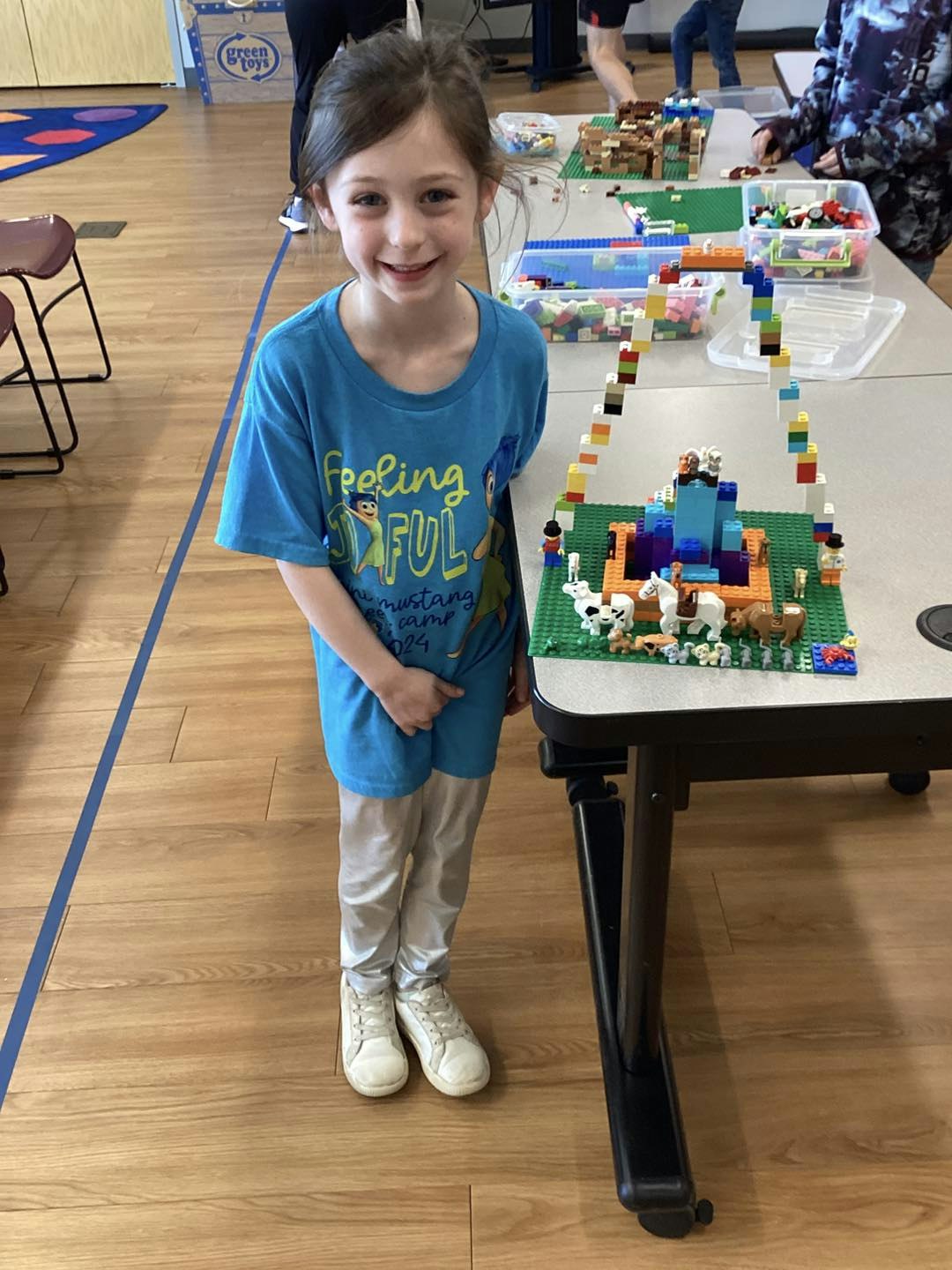 A child smiling beside a table with Lego structures, wearing a blue shirt.