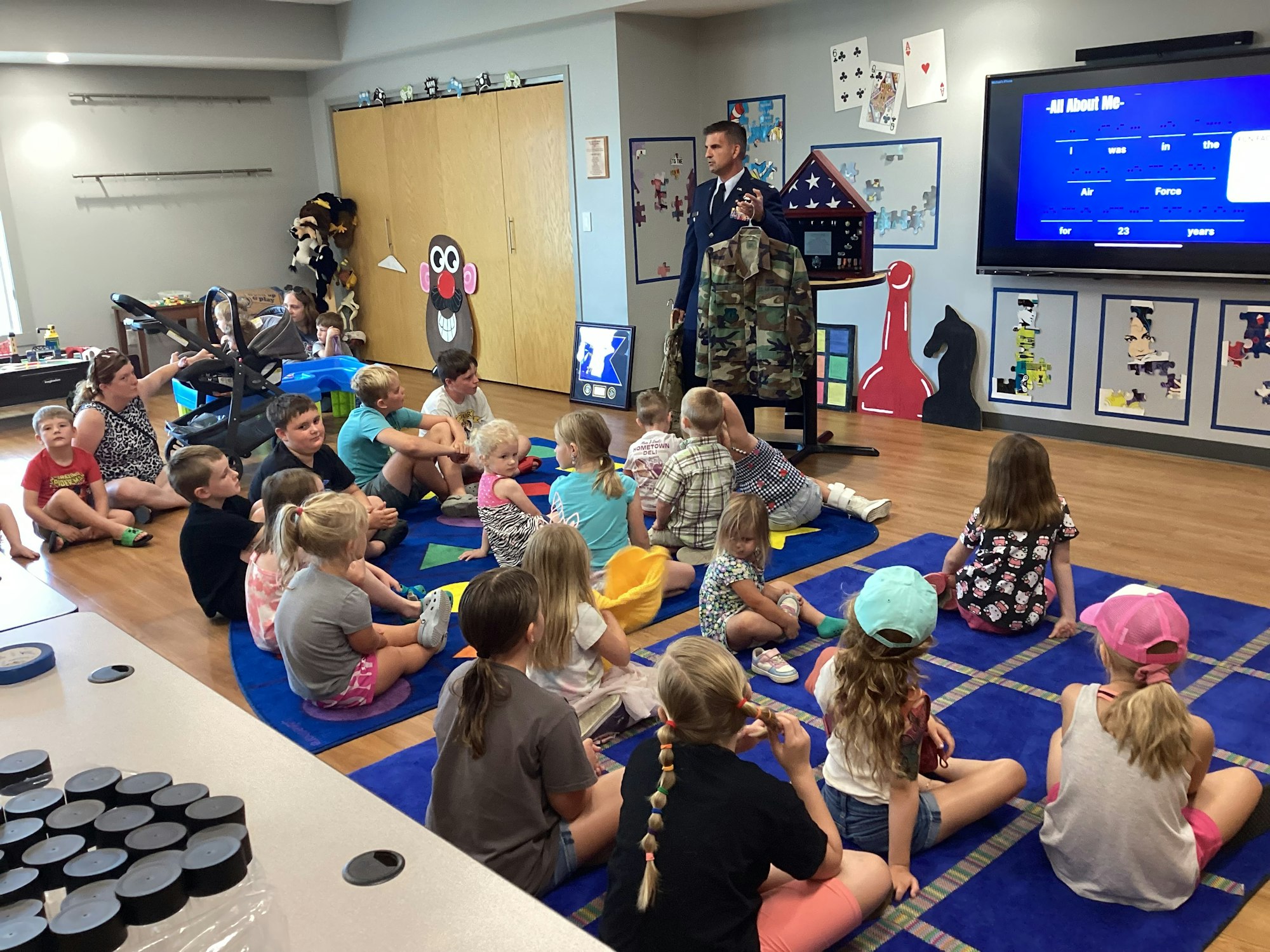 A person in uniform speaks to seated children in a classroom, using a digital screen for a presentation.