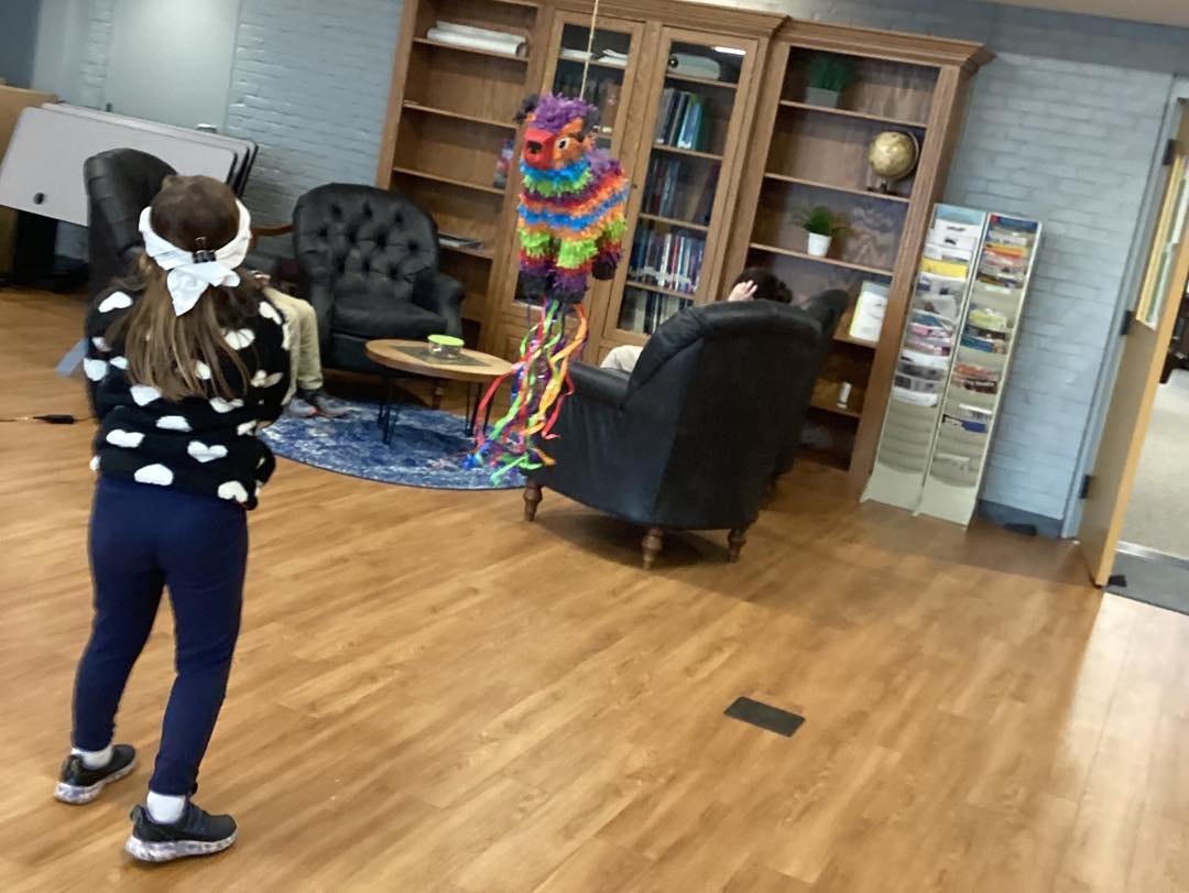 A child with a blindfold prepares to hit a colorful piñata in a room with people sitting and bookshelves in the background.