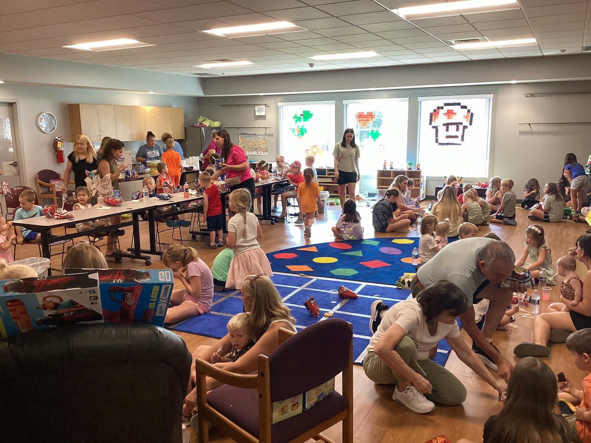 Children and adults in a room; tables with food, colorful rugs, and large windows with pixel art decorations.