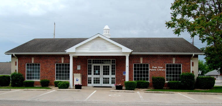 A small brick building with a peaked roof labeled "Four Star Public Library," surrounded by bushes and a parking area.