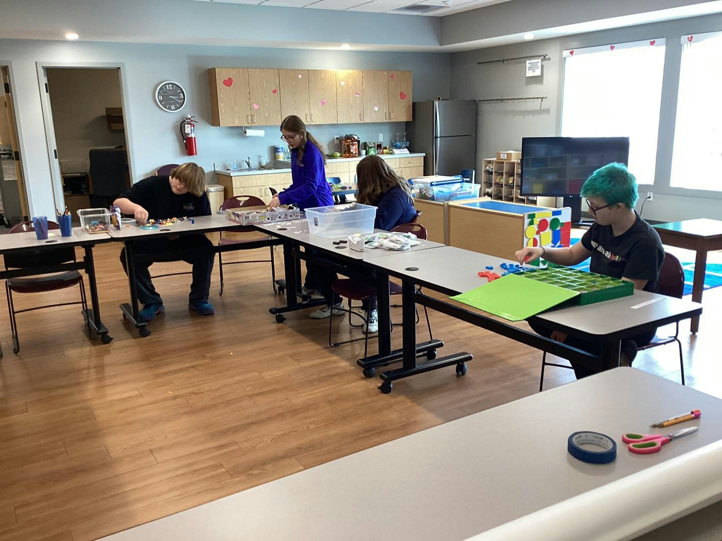 Four people are seated at a table in a room with craft supplies, working on various projects.