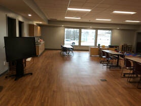A spacious room with wooden flooring, chairs, tables, a TV stand, and large windows.