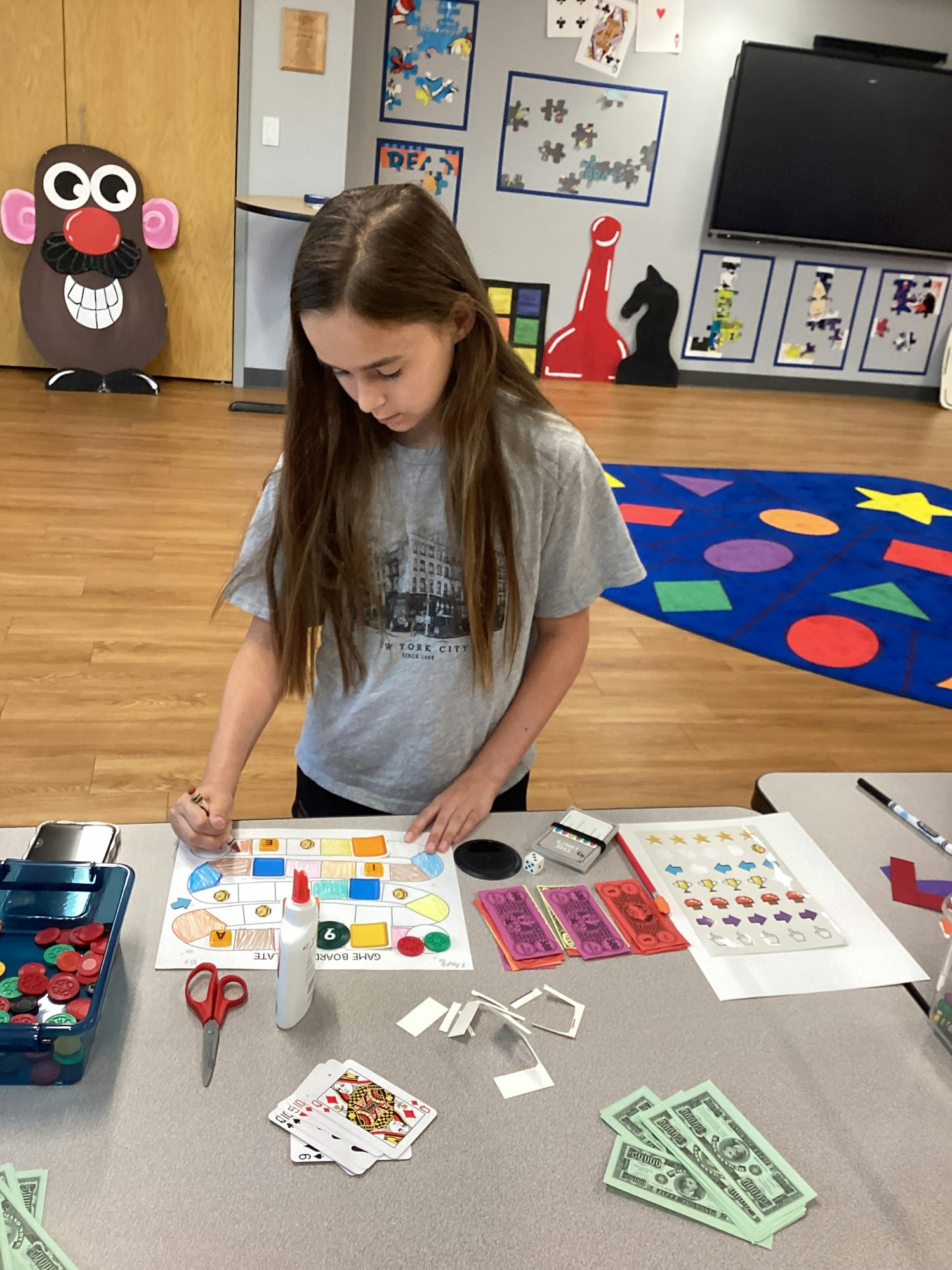 A person is working on a board game design in a colorful classroom with game pieces, cards, and play money on the table.