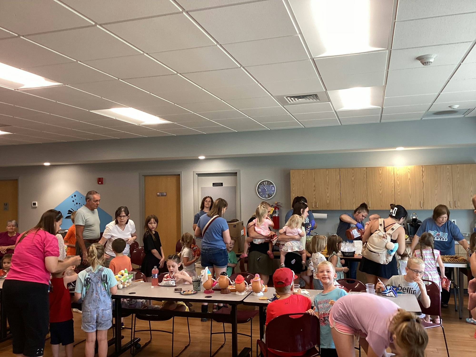 A group of people, including children, are gathered in a room with tables and chairs, engaging in activities.