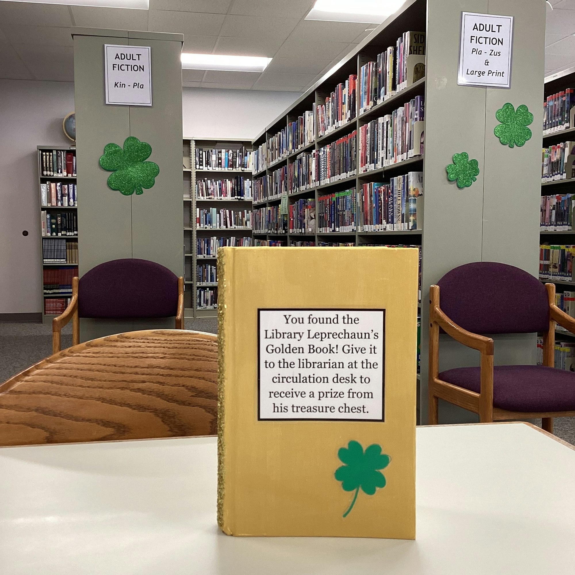 A golden book in a library with a sign offering a prize for finding it, surrounded by bookshelves and green shamrock decorations.