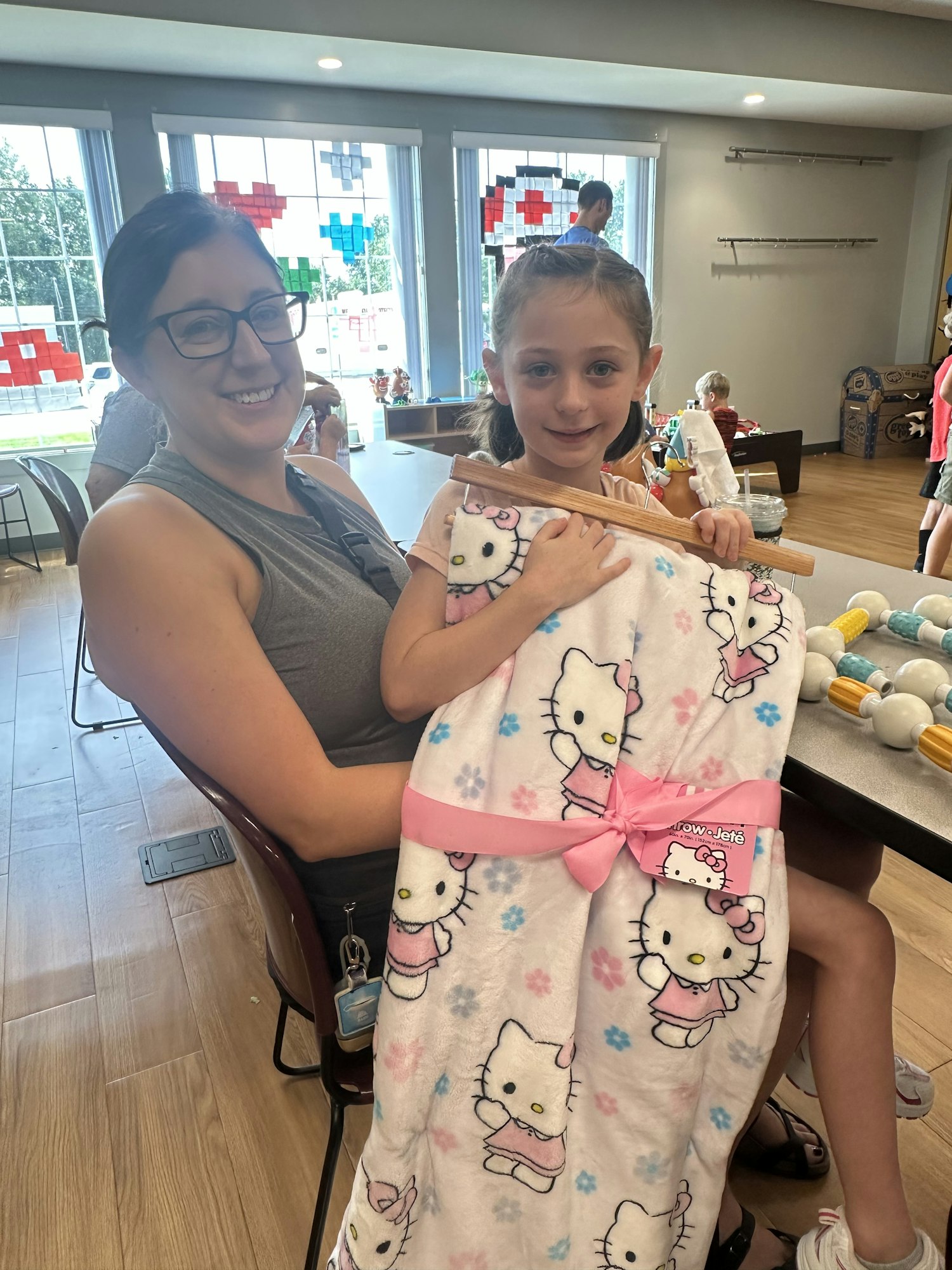 Girl holding a Hello Kitty blanket, sitting on a woman's lap in a brightly lit room with large windows.