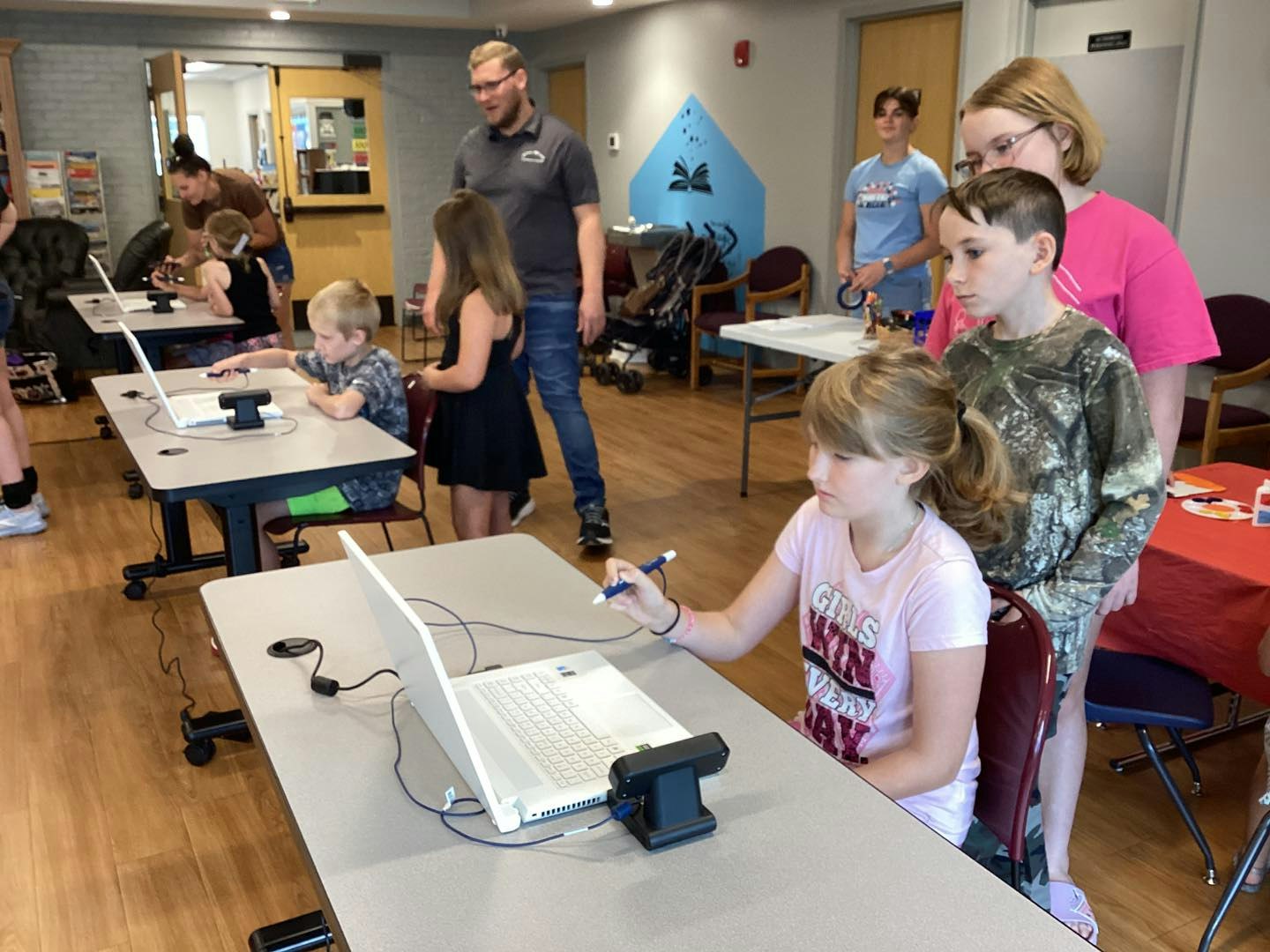 Children using laptops and VR controllers in a classroom or workshop setting, with adults overseeing.
