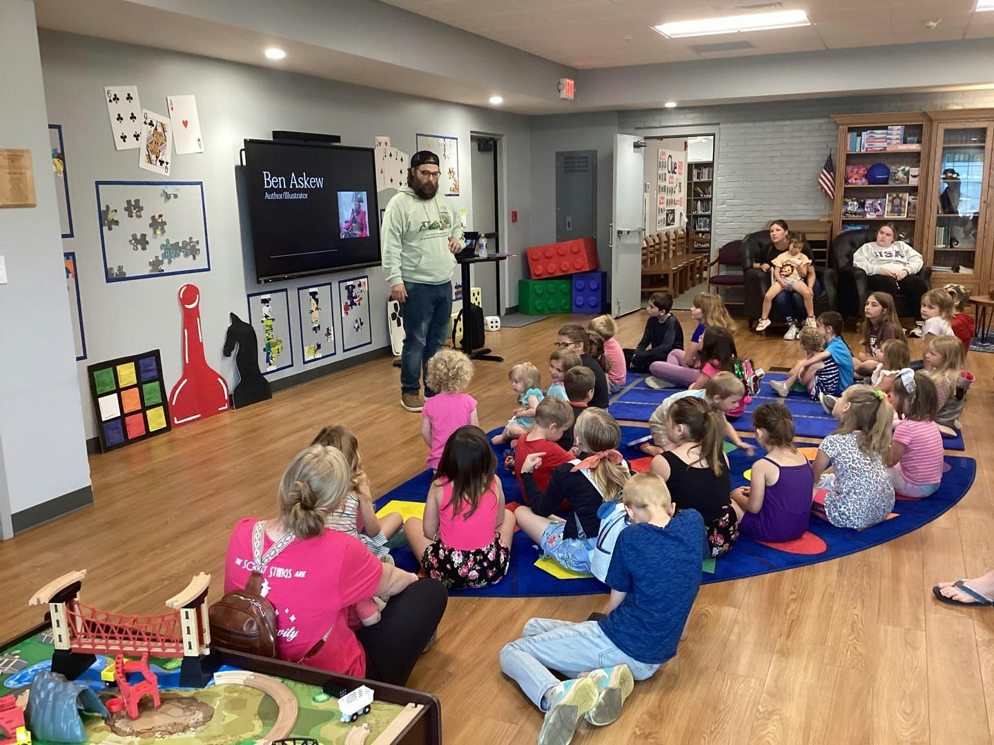 A man is speaking to a group of seated children in a colorful playroom with toys and wall decorations.