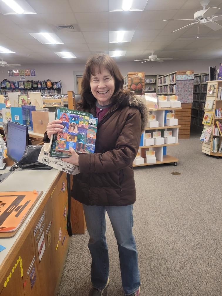 A smiling woman stands in a library holding a colorful book titled "Sticker by Number," with bookshelves in the background.