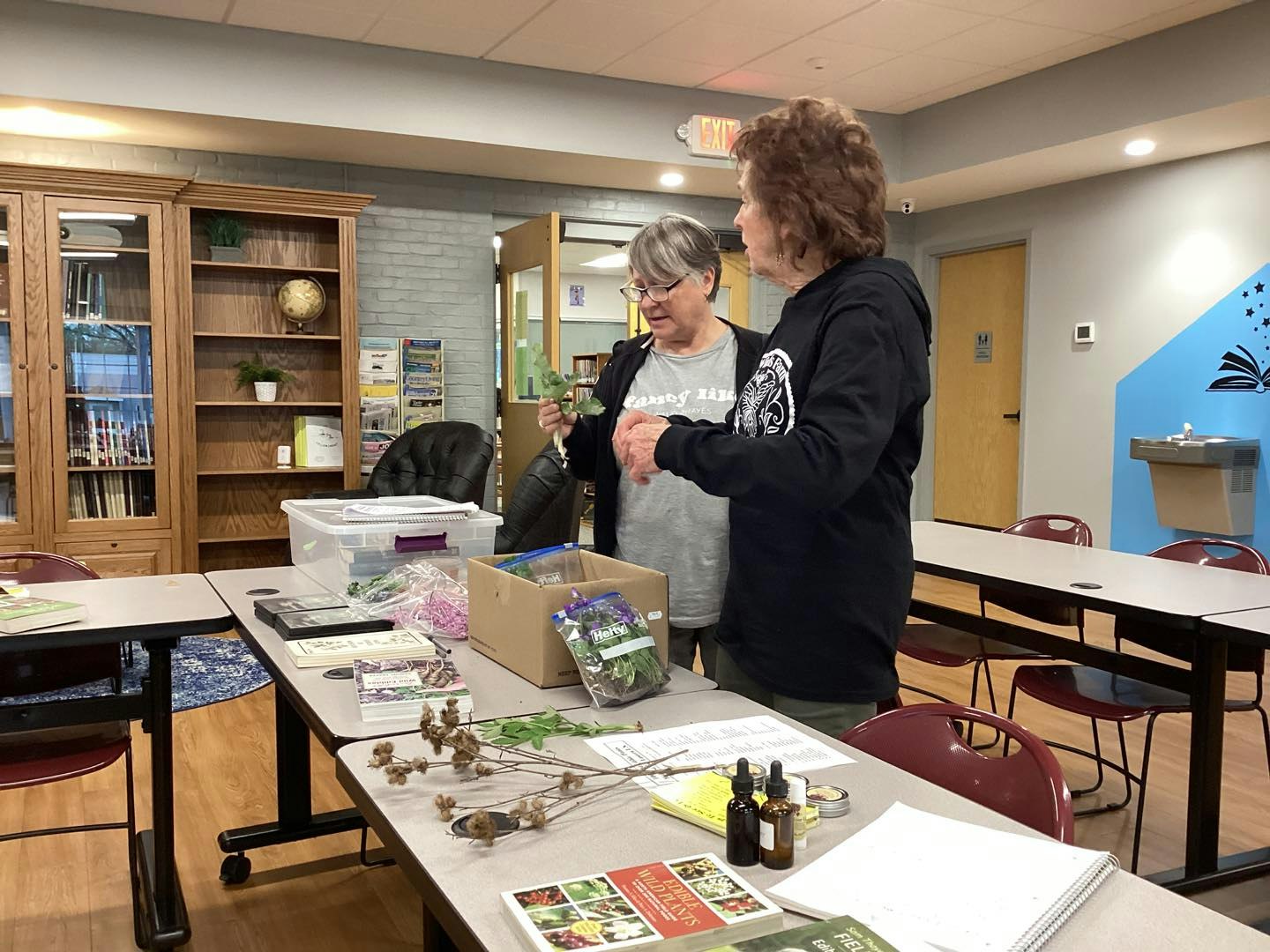 Two people examining plants at a table with books and materials in a room.