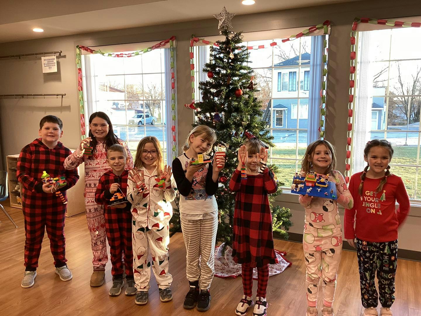 Children in pajamas smiling by a decorated Christmas tree, holding toys or crafts.
