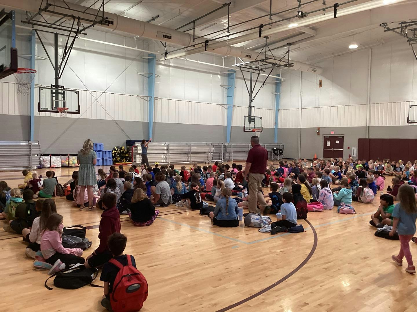 A large group of children sit on a gym floor with backpacks, while adults stand nearby, likely in a school assembly.