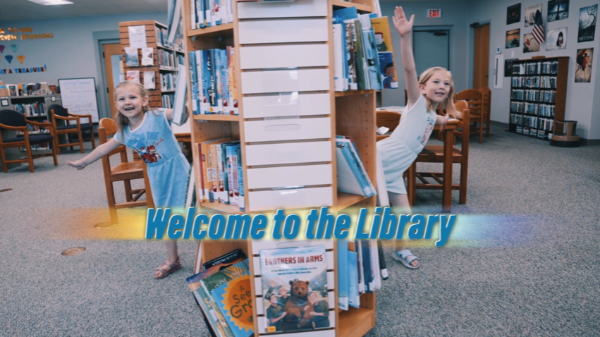 Two children in a library, smiling and waving near shelves. Text: "Welcome to the Library."