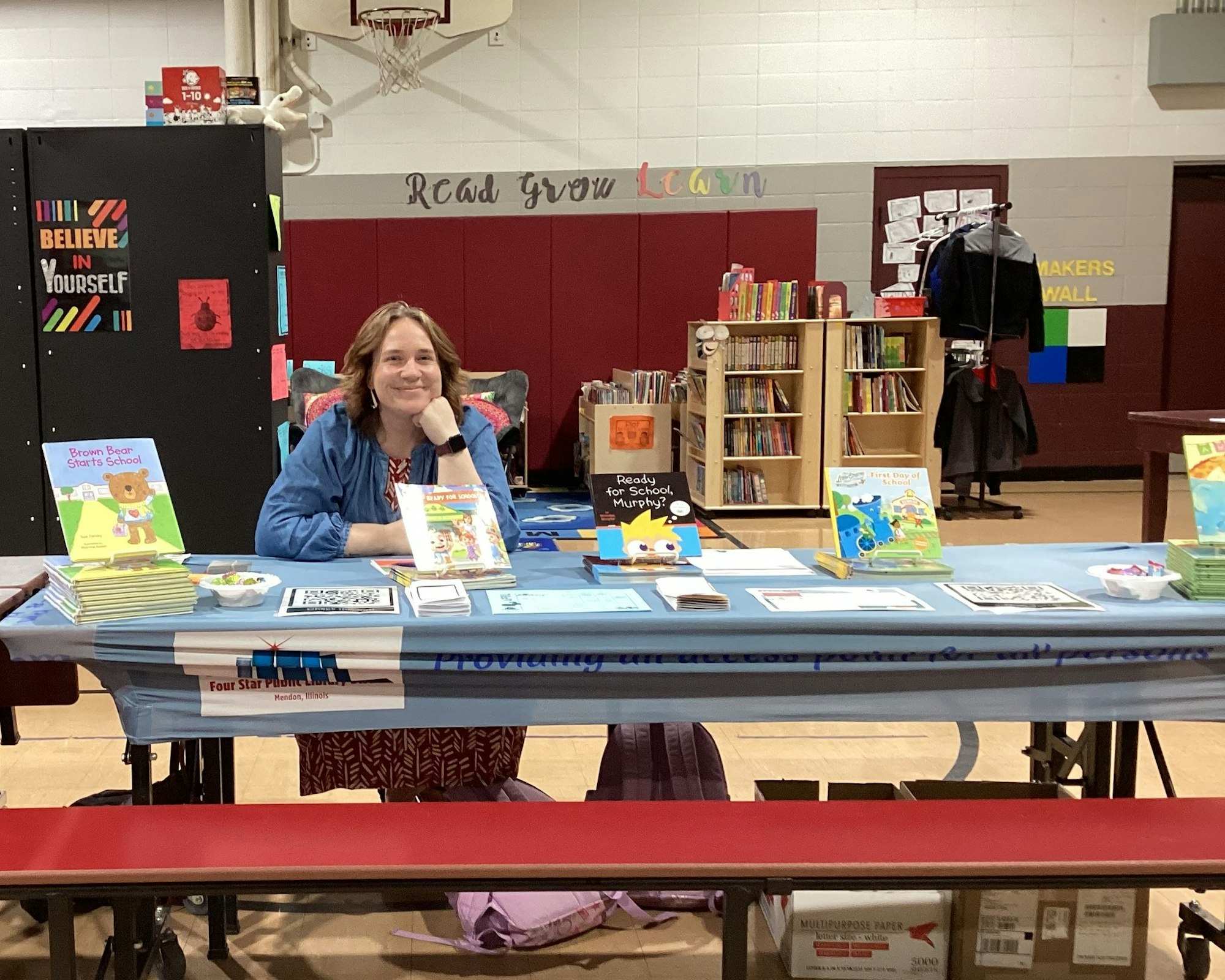 A person sits at a table with children’s books and promotional materials, in a gym-like setting.