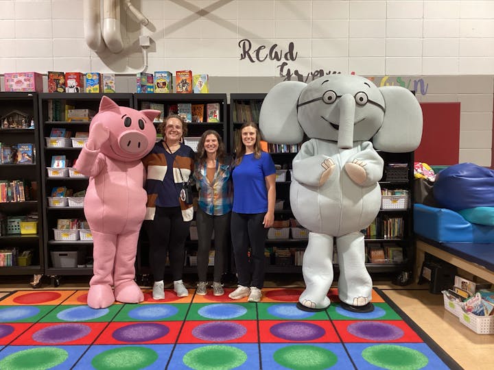 Two people in pig and elephant costumes stand with three others in a colorful library setting.