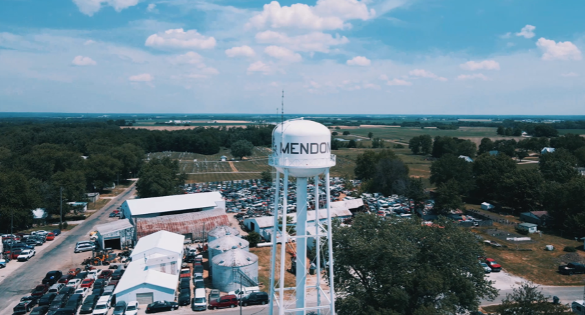 A water tower labeled "MENDON," surrounded by buildings, trees, and a parking lot filled with cars under a blue sky.