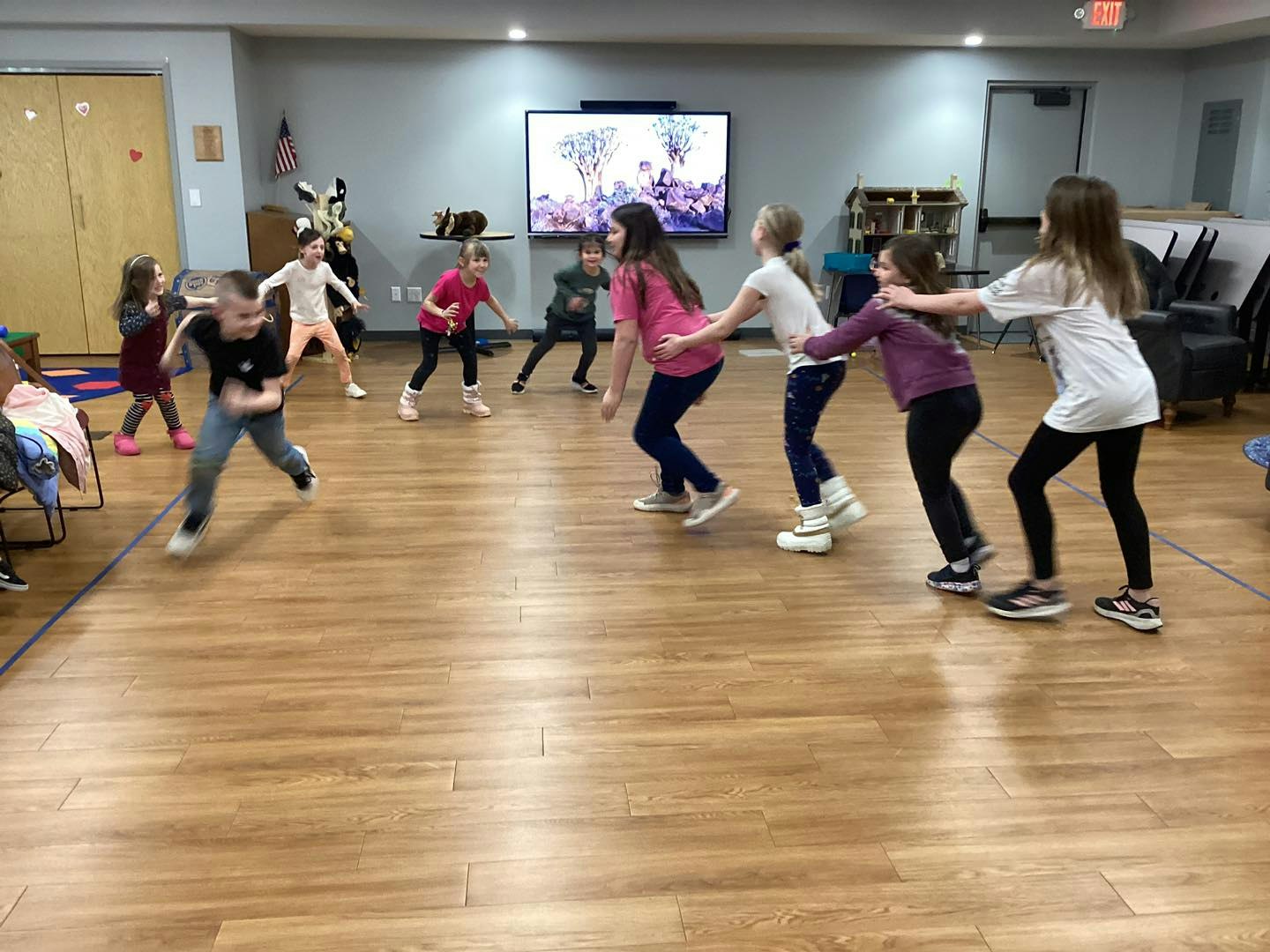 Children playing a game inside a room with wood flooring, forming a line and moving in a circle.