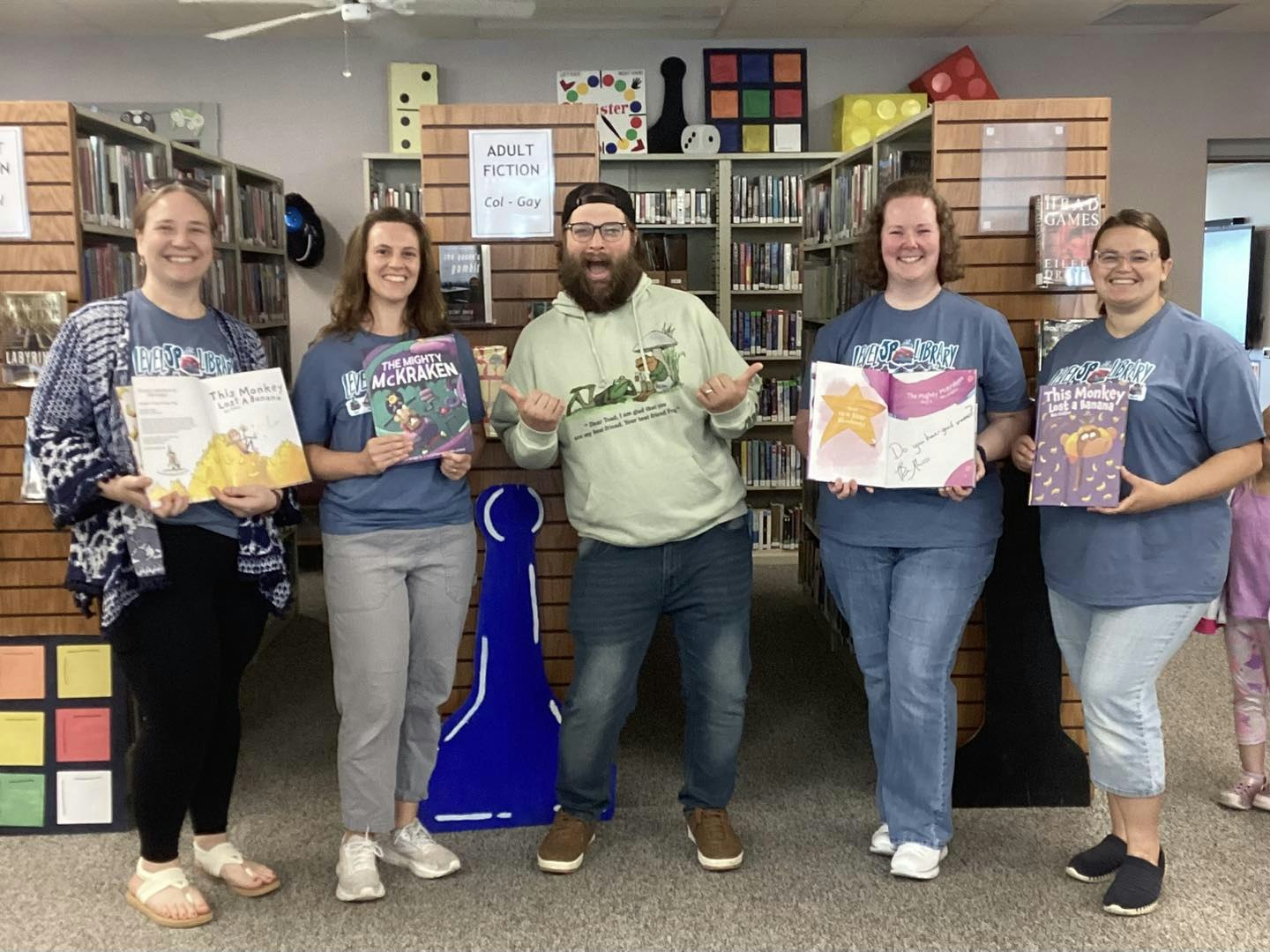 Five people in a library hold children's books, smiling. Shelves of books are in the background with signs for adult fiction.