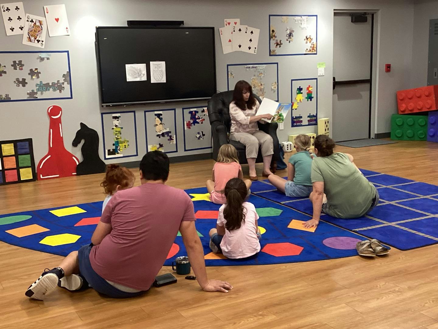 A woman reads to children and adults seated on a colorful rug in a classroom with large playing cards and puzzle decor on the walls.