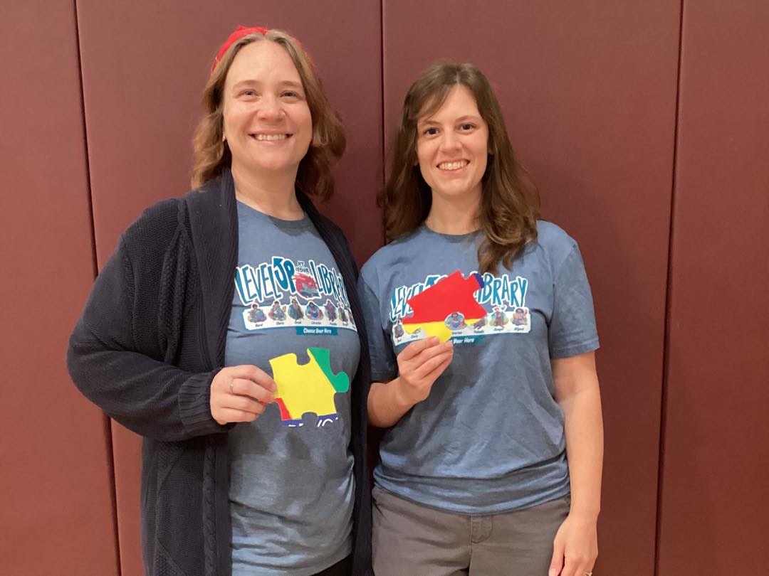 Two people smiling, wearing matching t-shirts, holding colorful puzzle pieces against a maroon background.