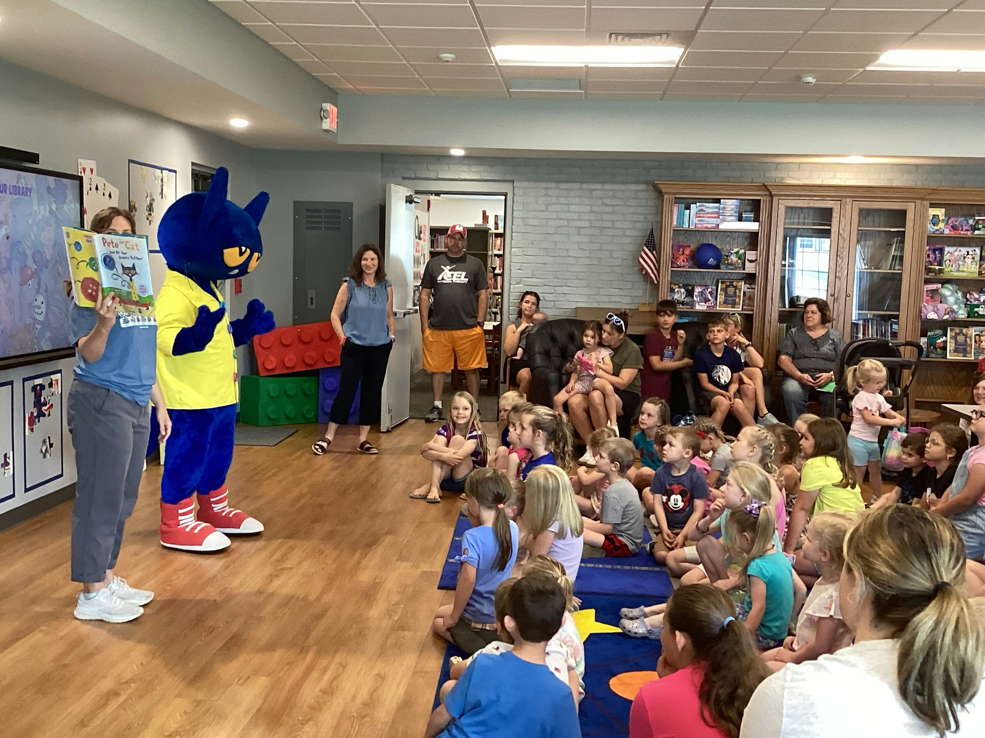 A person in a Pete the Cat costume and a woman hold a "Pete the Cat" book, surrounded by children and adults in a library setting.