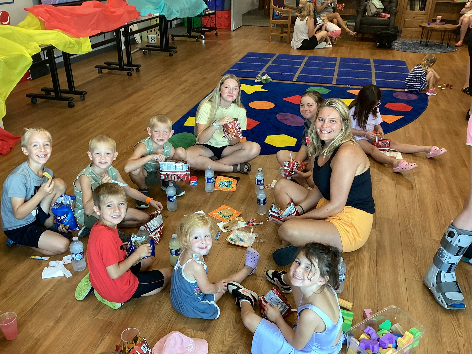 A group of kids and an adult are sitting on the floor with snacks and drinks, smiling for the camera.