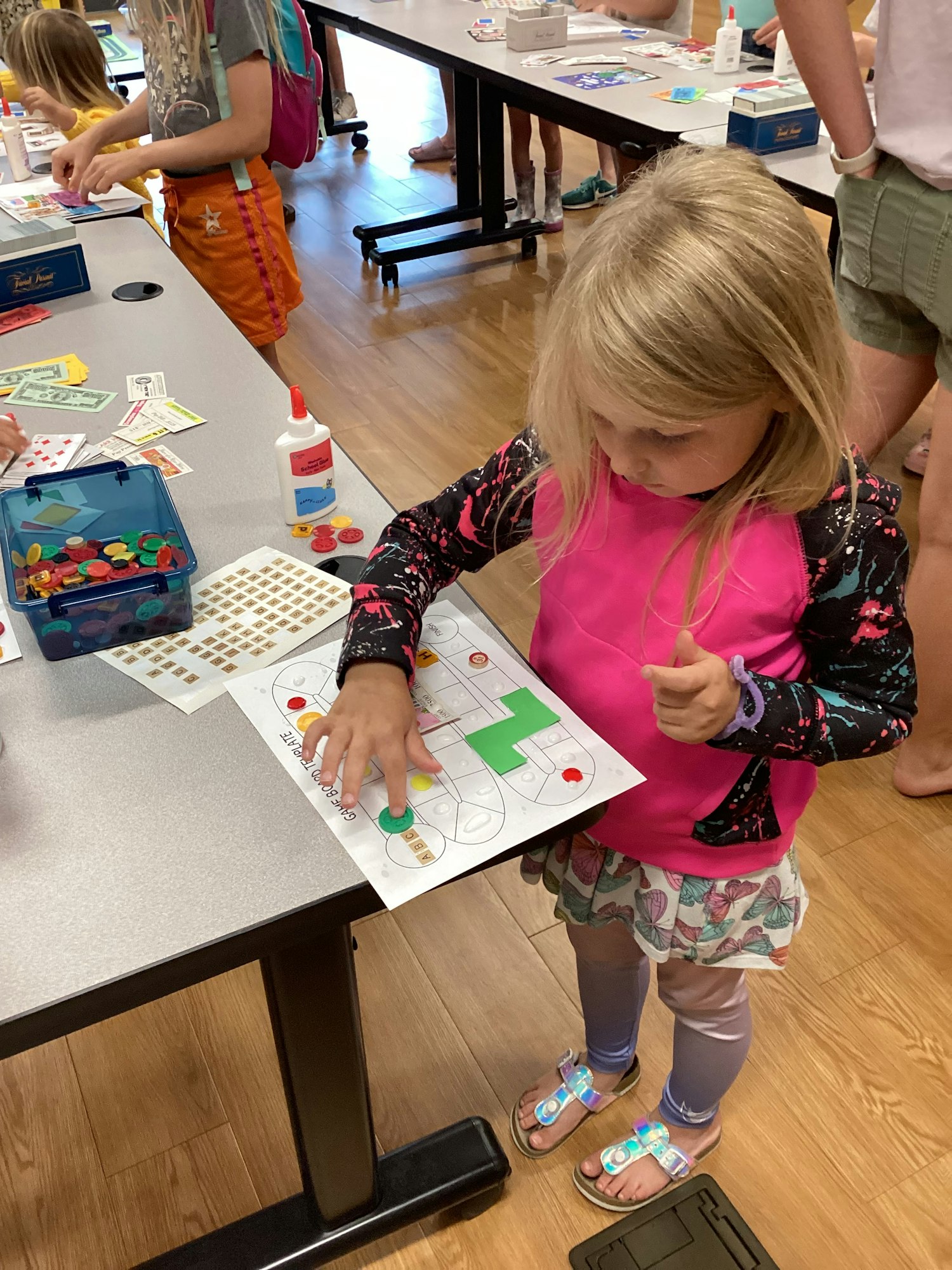 Child playing a board game on paper at a table with art supplies and other children around.