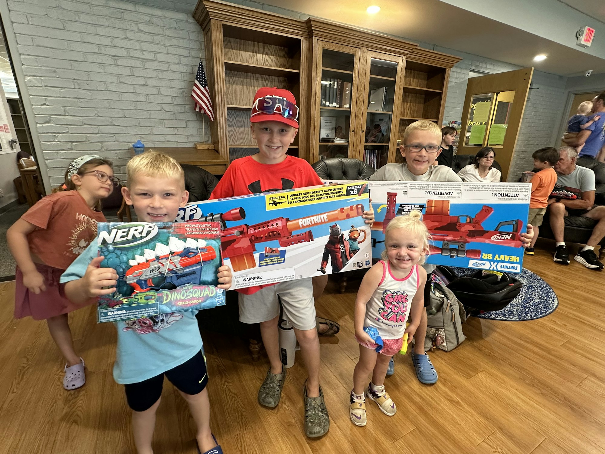 Kids holding Nerf guns in a room with people sitting in the background.