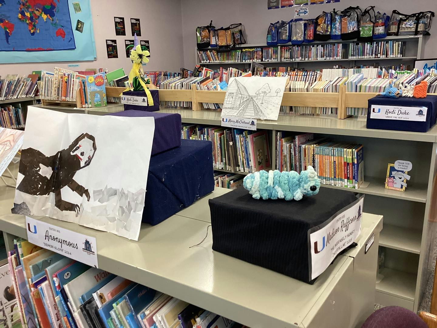 Library shelves with books and art projects displayed, including drawings and a plush caterpillar.