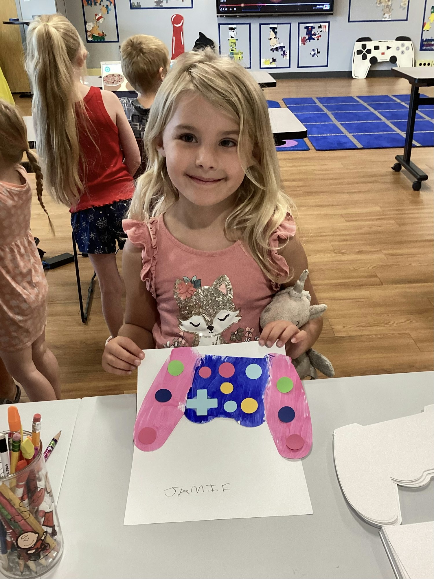 A child in a classroom shows a decorated paper game controller.