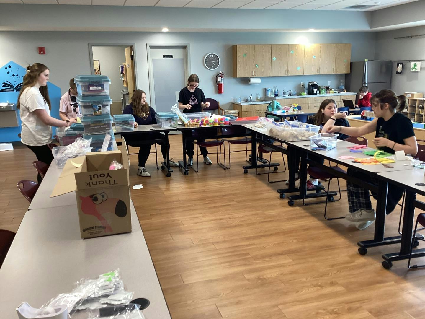 A group of people in a room are organizing containers and crafting materials on tables.