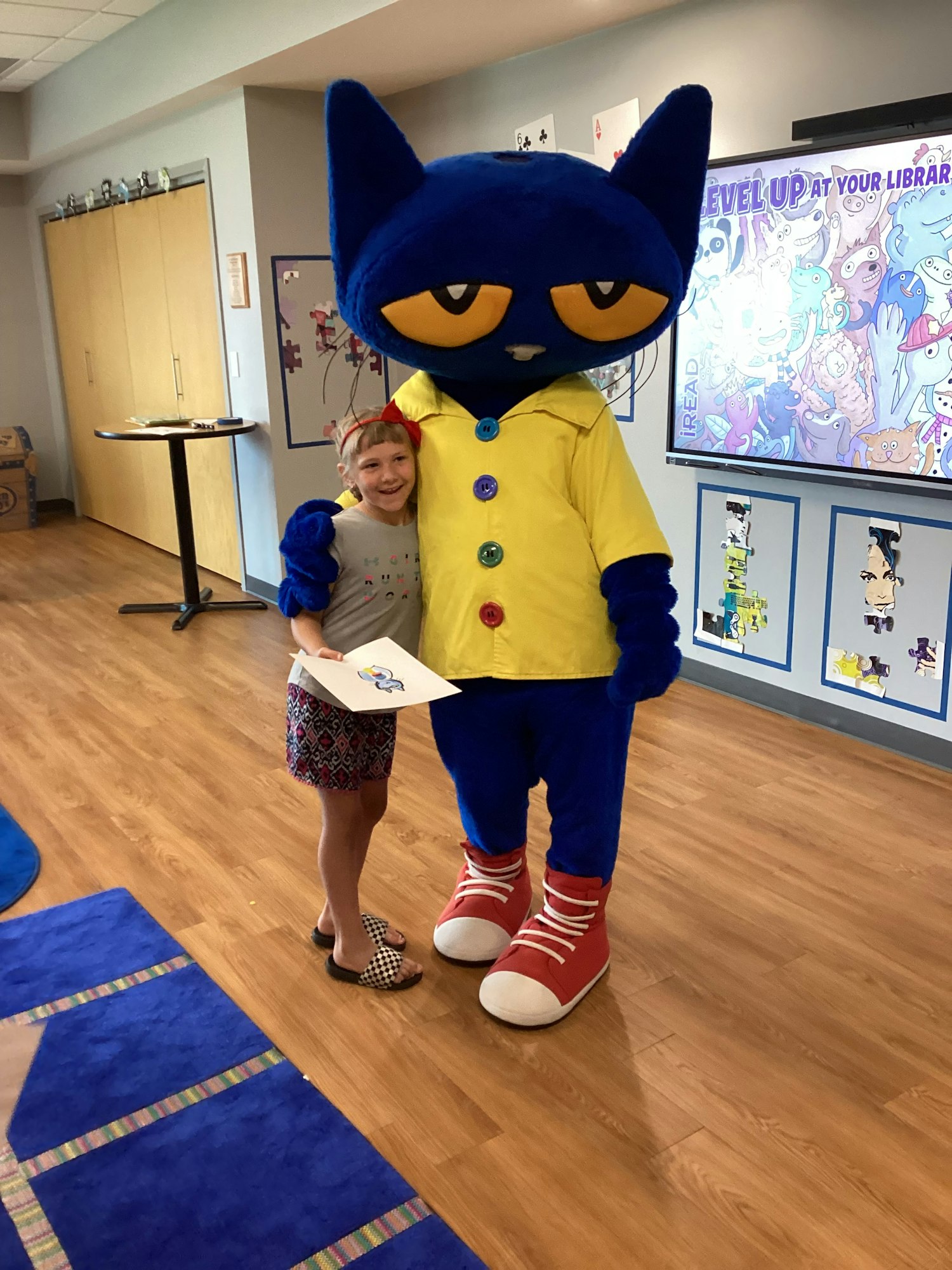 A child stands with a person in a large blue cat costume inside a room with colorful decorations.