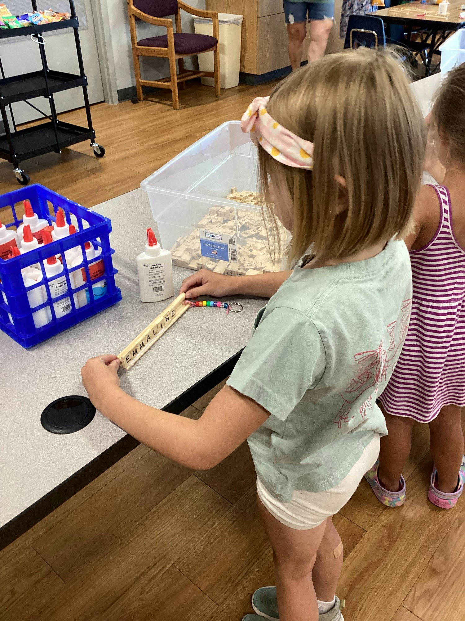Children at a table with a glue bottle, plastic container of tiles, and a rack of glue bottles.