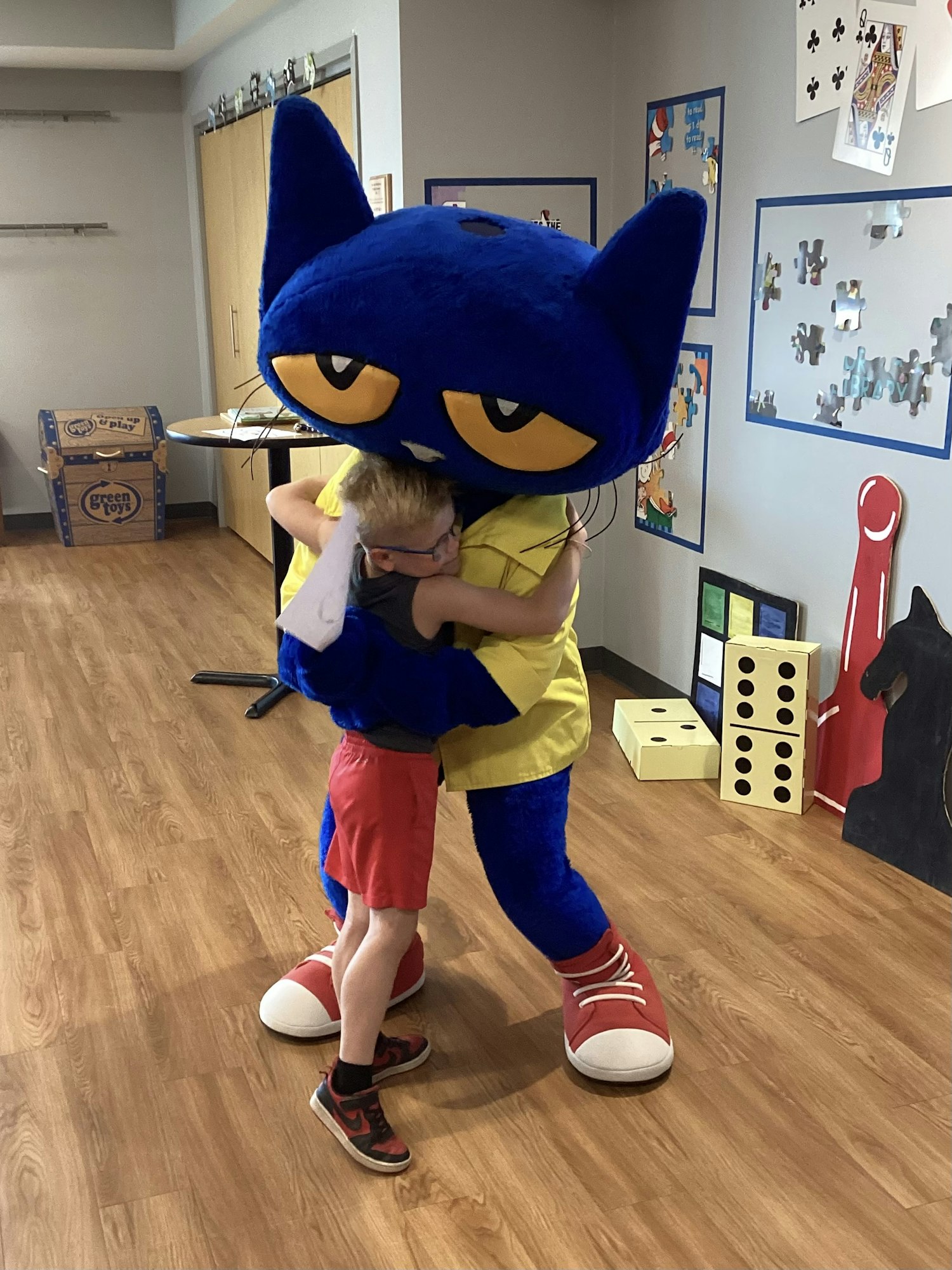 A child hugs a large mascot costume of a blue cat in a room with educational decorations.