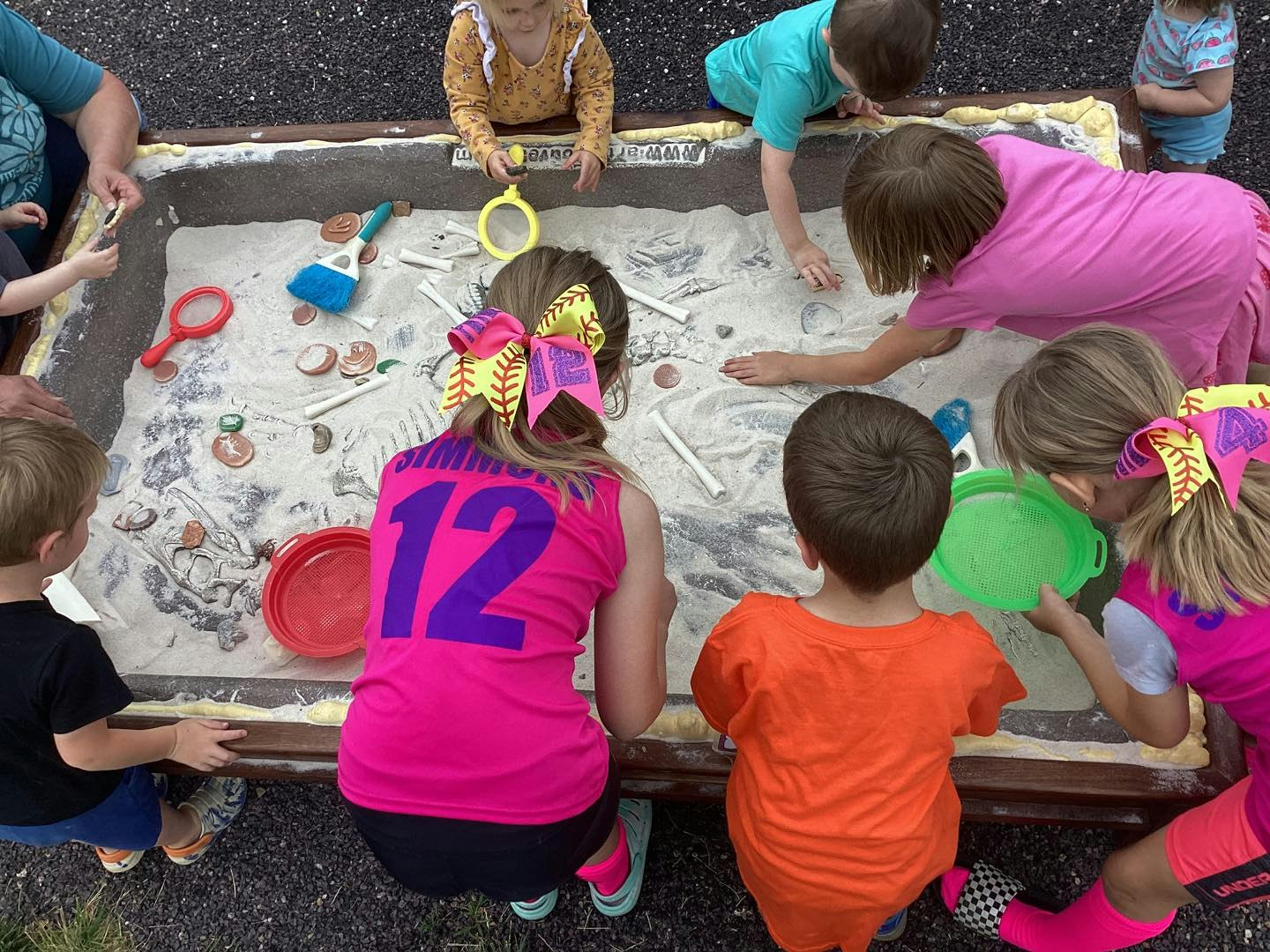 Children playing in a sand table with dinosaur bones and tools for digging.