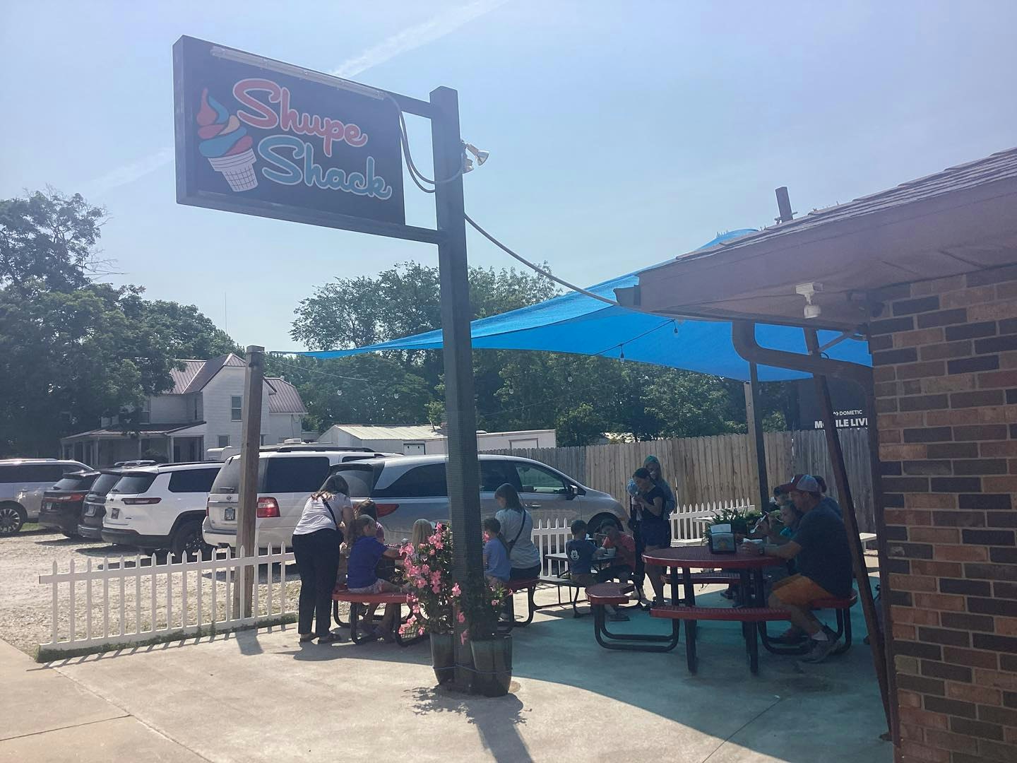 Outdoor seating at "Shupe Shack" with people enjoying under a blue canopy, cars parked nearby.
