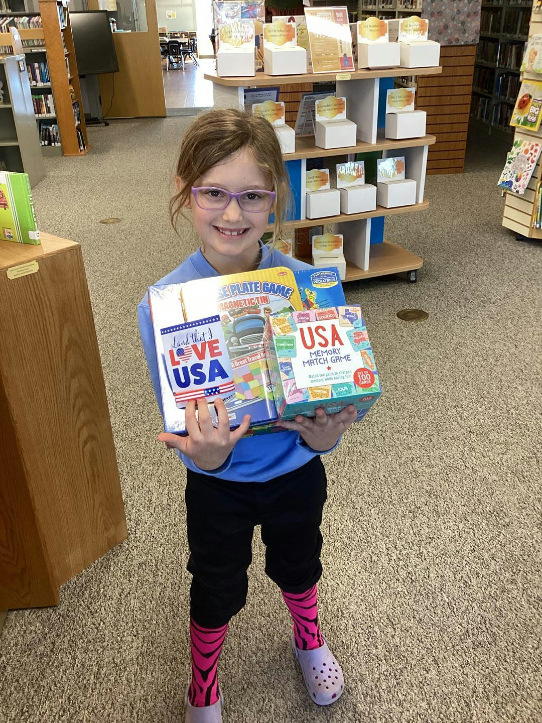 A child in a library holds games themed around the USA, smiling while wearing colorful socks and glasses.