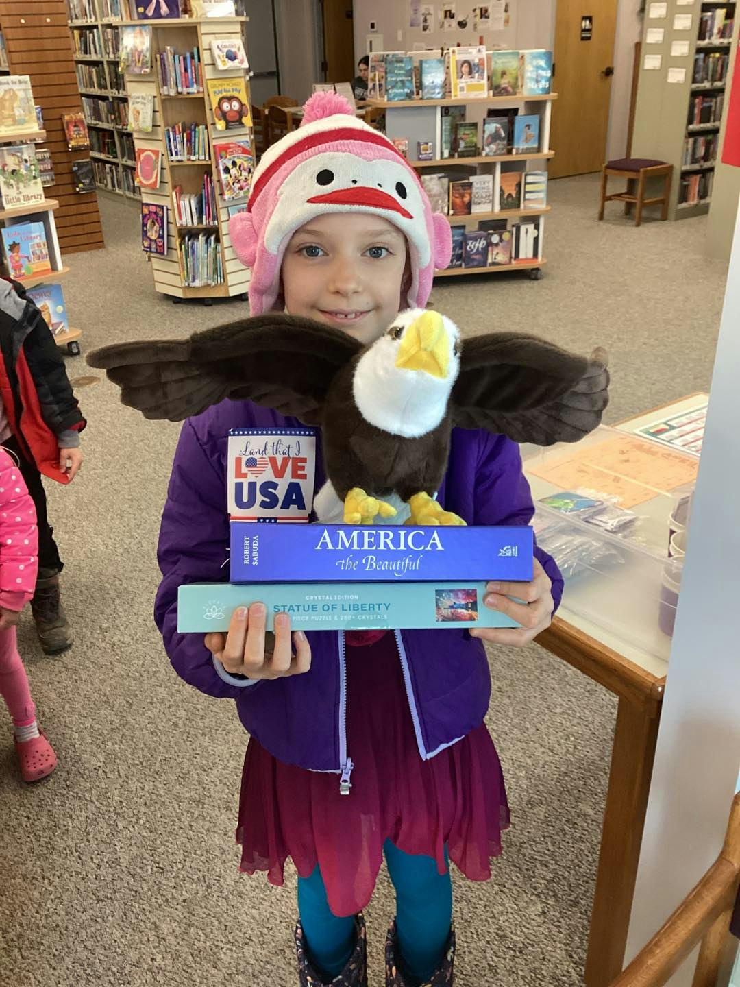A girl in a colorful outfit holds a plush eagle and books about America in a library, wearing a fun monkey hat.