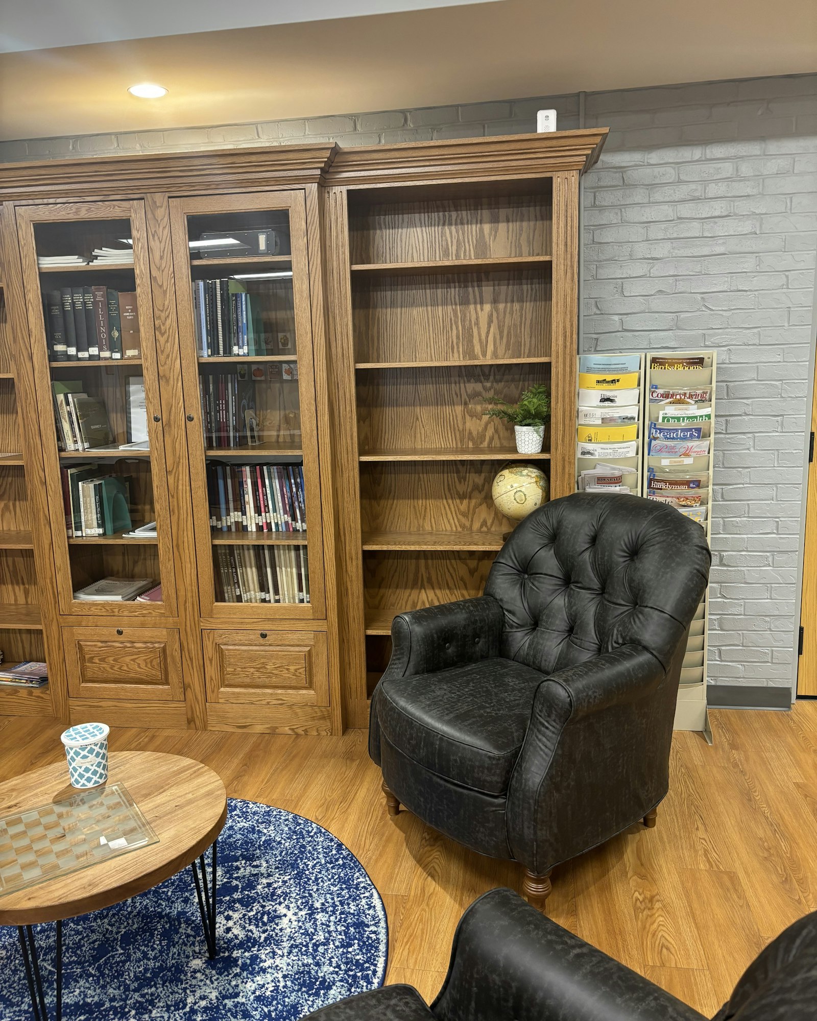 Cozy reading nook with armchair, wooden bookshelves, a globe, magazines, and a round table with a blue rug.